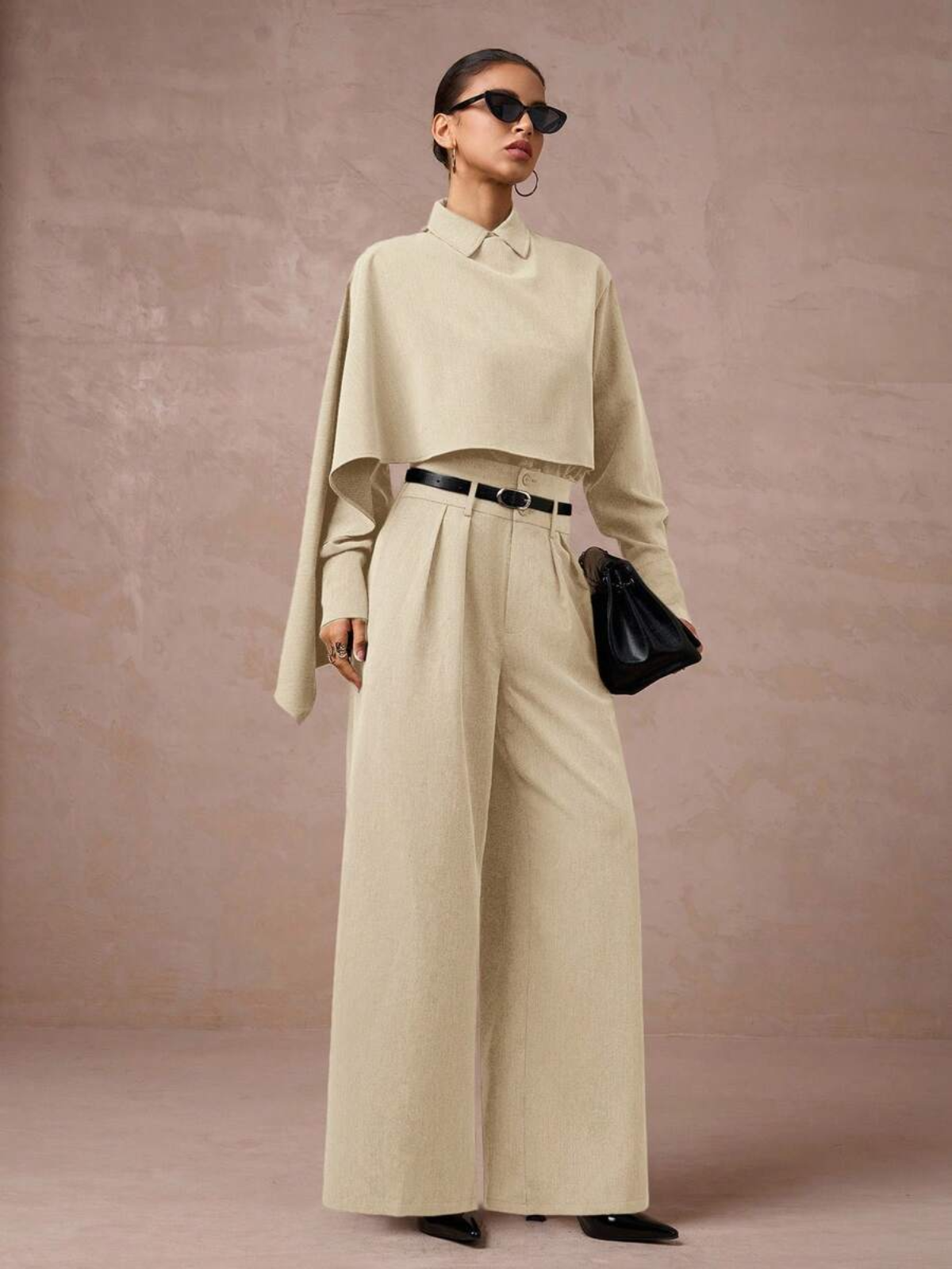 Completo beige elegante con blazer cropped e pantaloni larghi, cintura nera in vita, clutch nera e occhiali da sole.

