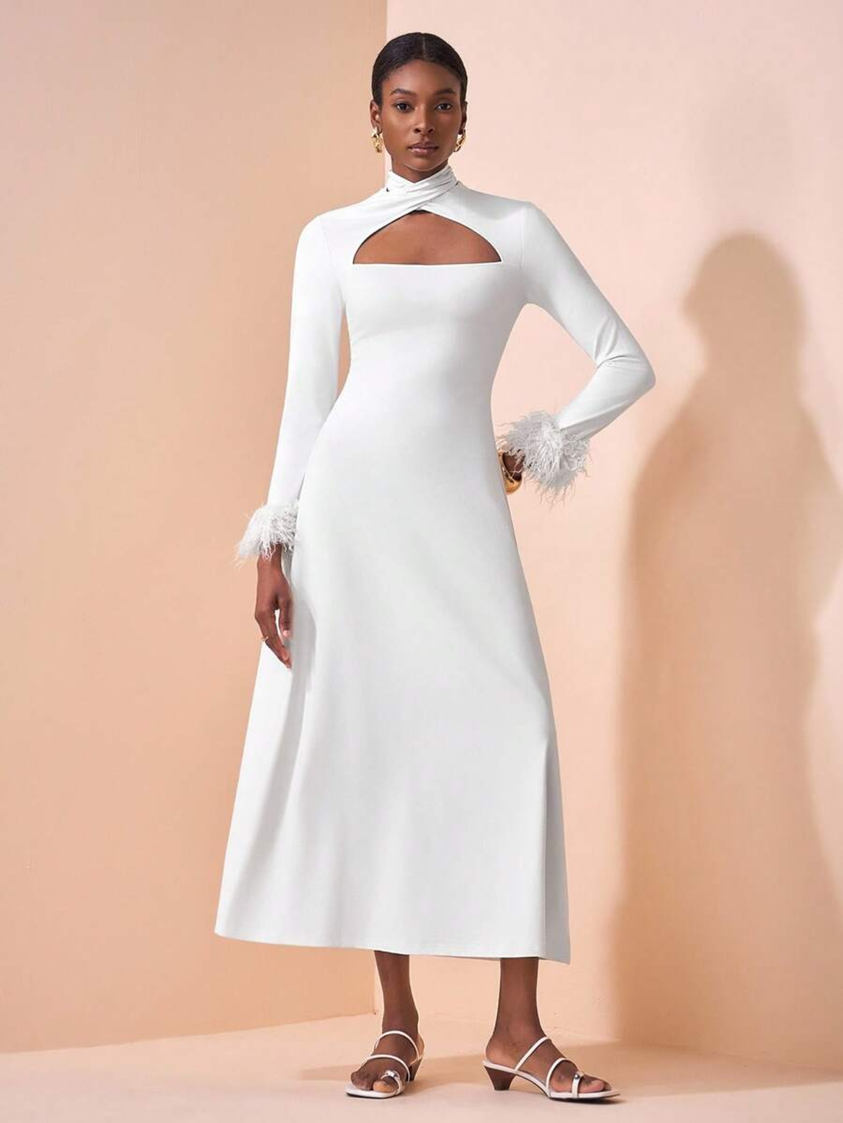 Donna che indossa abito lungo elegante con cut-out al petto, collo alto, maniche lunghe con dettagli in piume ai polsi, su sfondo rosa pesca.

