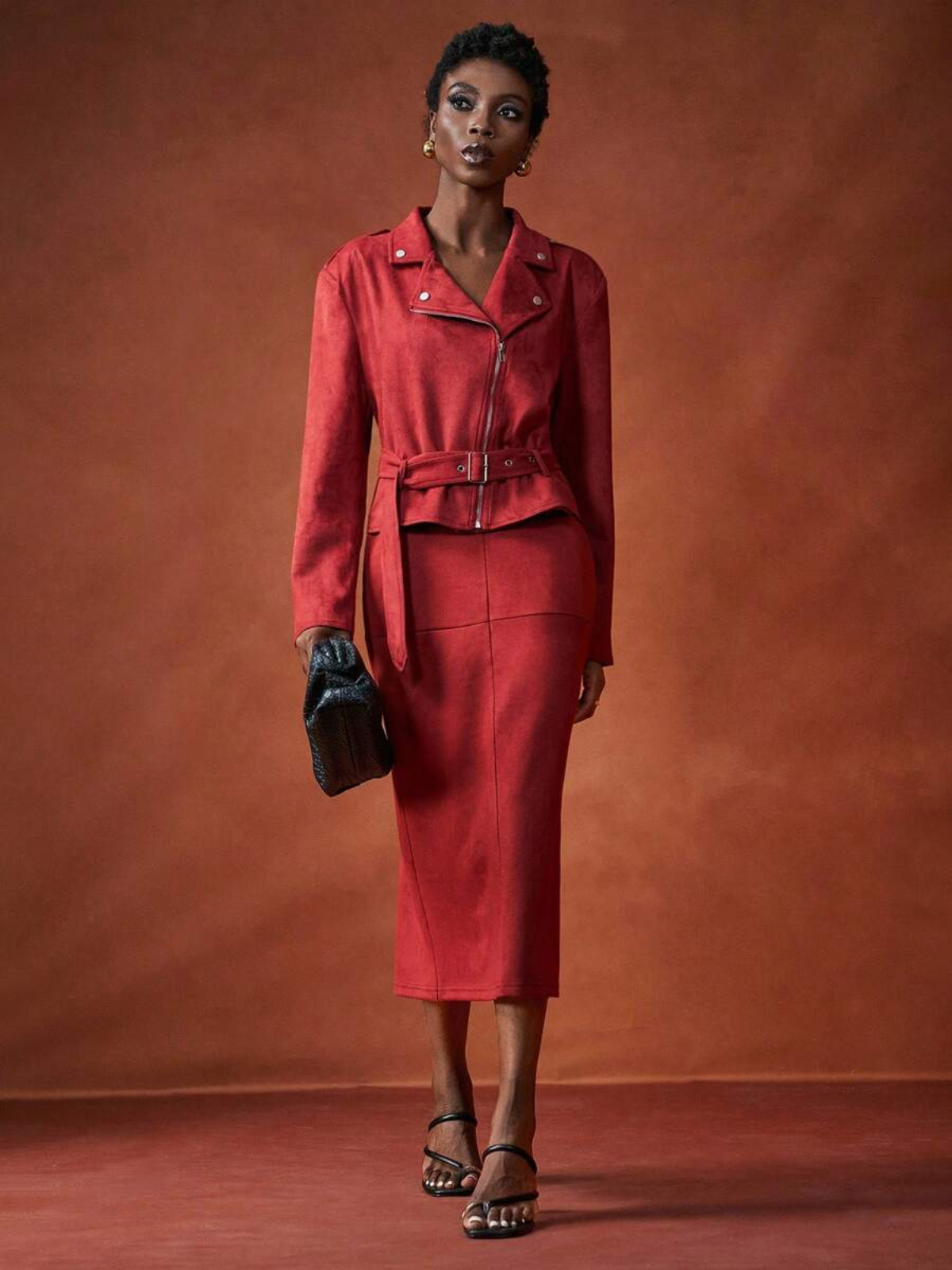 Completo rosso elegante con giacca biker chiusa e cinturata, gonna midi coordinata, clutch nera e sandali con tacco, look sofisticato.
