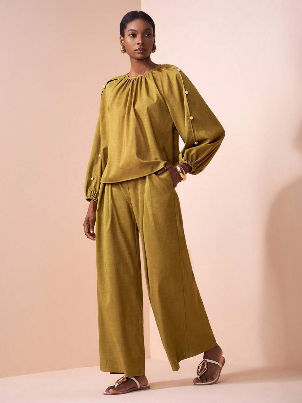 Donna che indossa completo senape con blusa a maniche lunghe e bottoni perla, pantaloni palazzo larghi, sandali cuoio su sfondo rosa.
