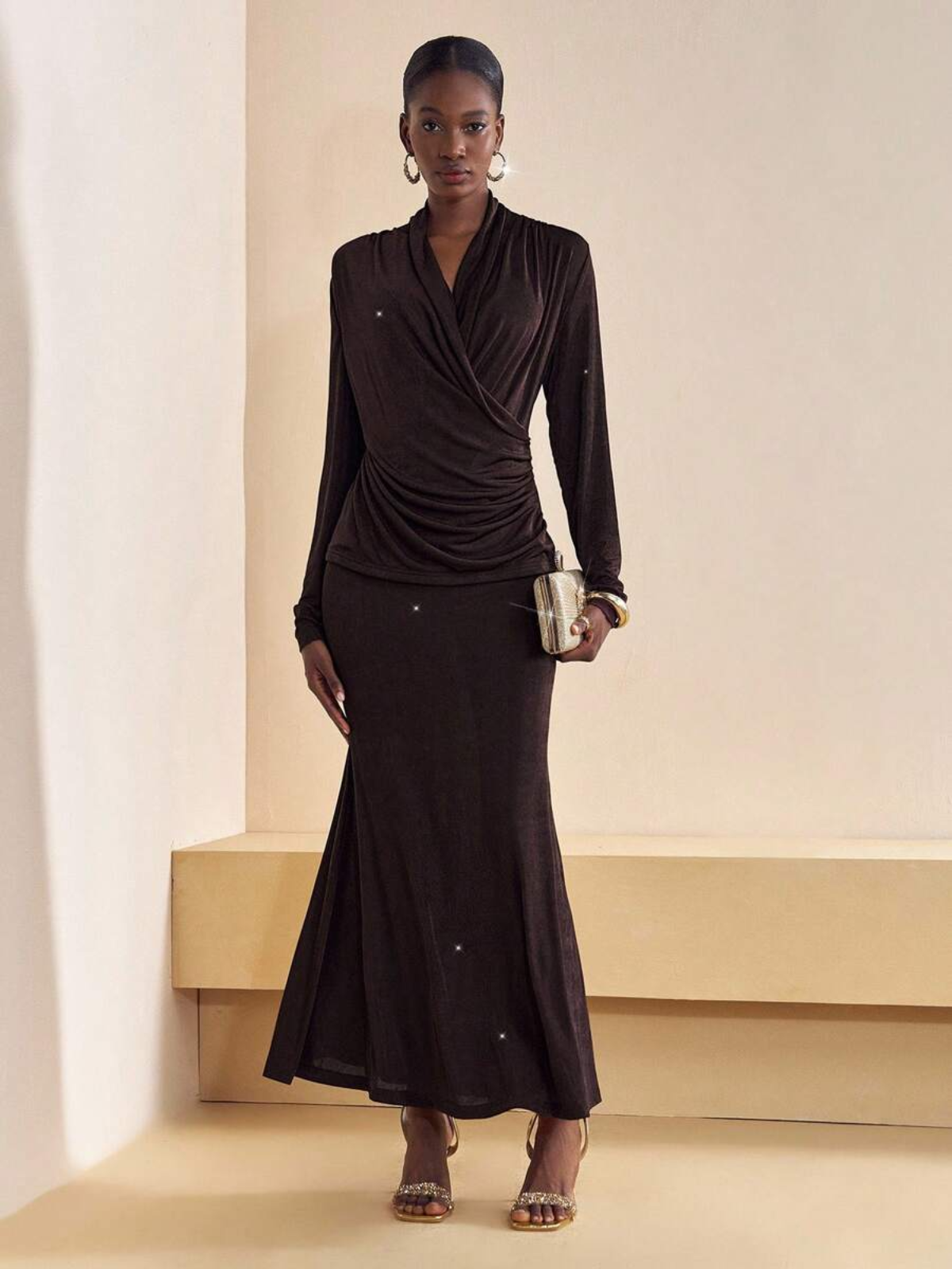 Abito marrone elegante con top avvolgente e gonna midi, modella con clutch dorata e sandali gioiello, look da sera sofisticato.
